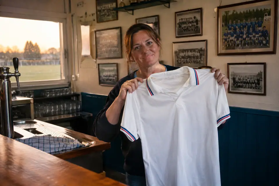 Une femme tient un maillot de foot blanc rétro brillant au comptoir d'un club amateur
