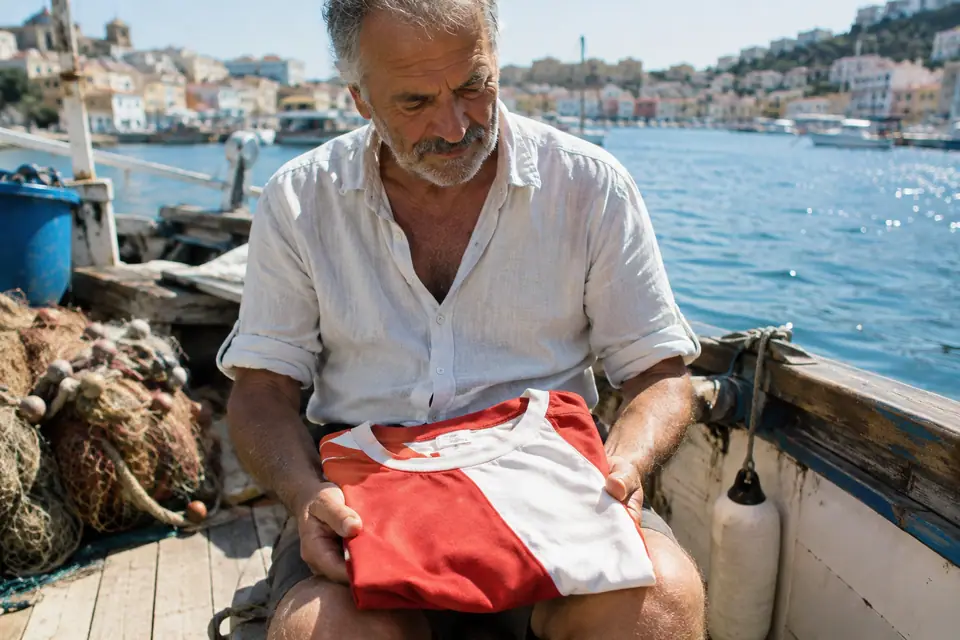 Homme tenant sur ses genoux un maillot de foot rétro partagé en diagonale rouge et blanche sur un bateau
