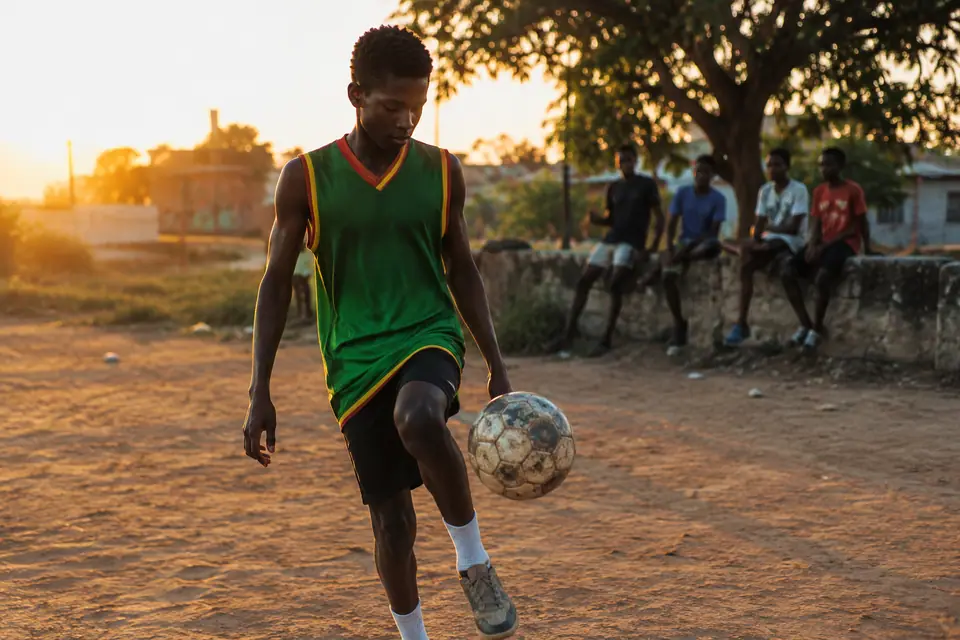 Un adolescent en maillot de foot vert sans manches jongle sur un terrain poussiéreux au coucher du soleil