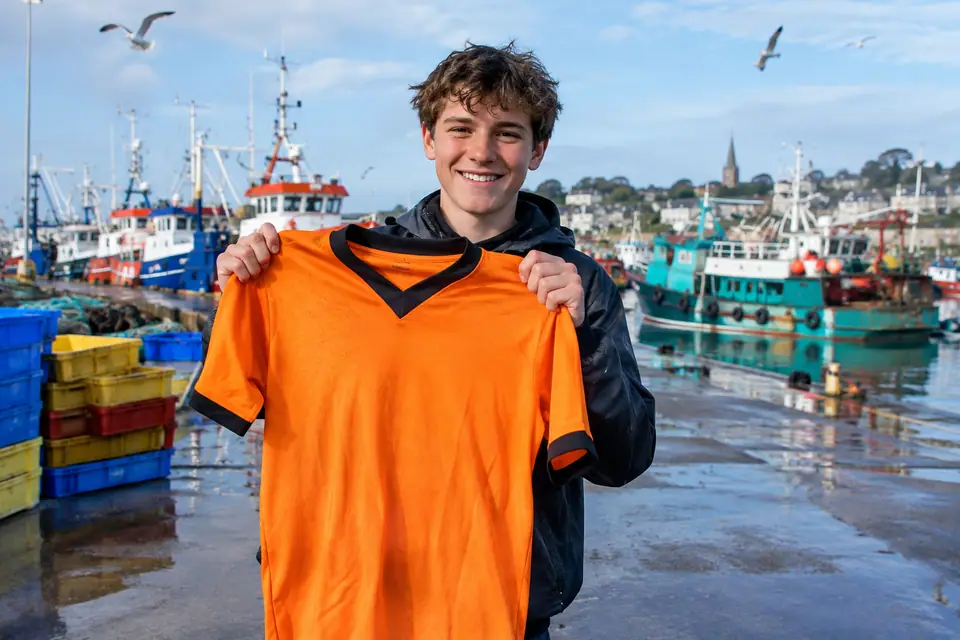 Adolescent tenant un maillot de foot orange à col noir sur le quai d'un port de pêche breton
