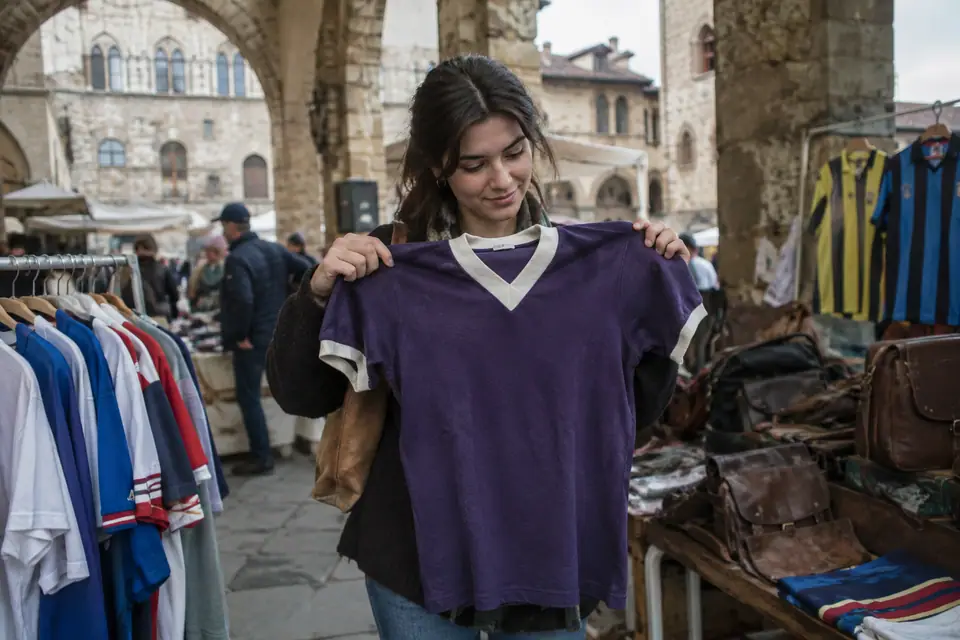 Jeune femme tenant un maillot de foot violet rétro sur un stand de brocante sous des arcades en Toscane