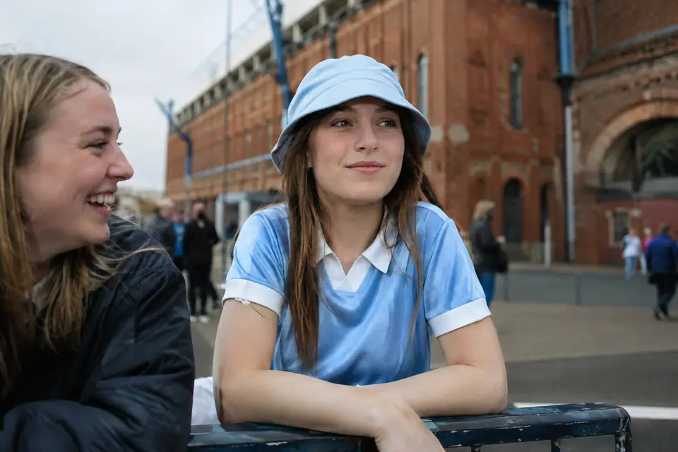 Adolescente en maillot de foot bleu ciel rétro et bob, appuyée sur une barrière devant un vieux stade de brique