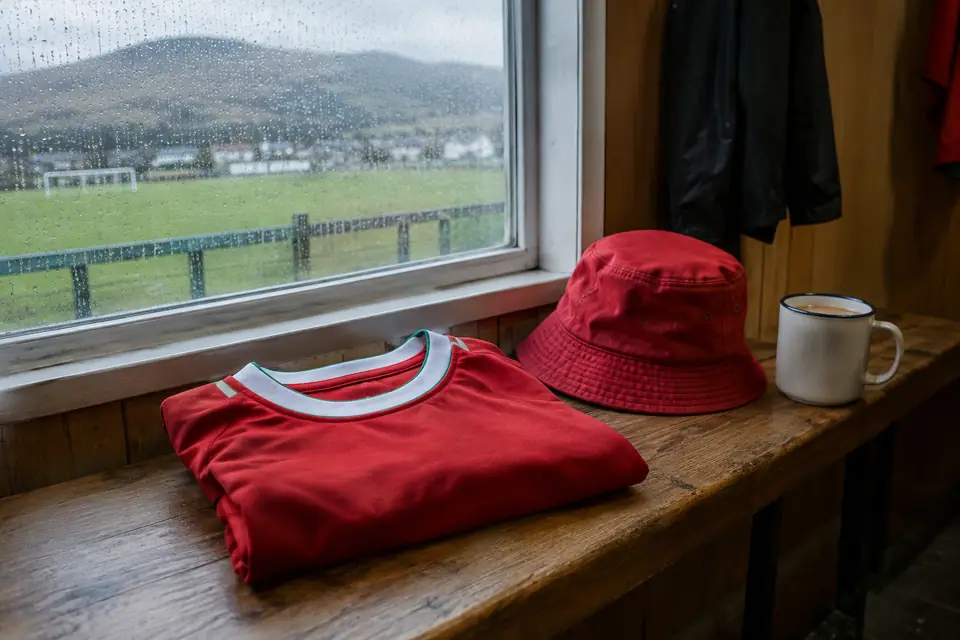 Maillot de foot rouge rétro plié sur un banc de club house, avec un bob et une tasse de thé