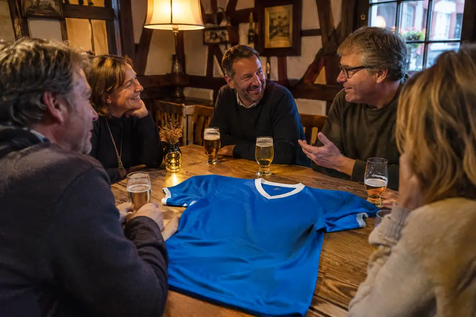 Maillot de foot rétro bleu à col blanc posé sur une table de winstub entouré d'amis