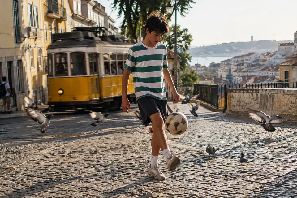 Un adolescent en maillot de foot rétro à rayures horizontales vertes et blanches jongle sur une place pavée