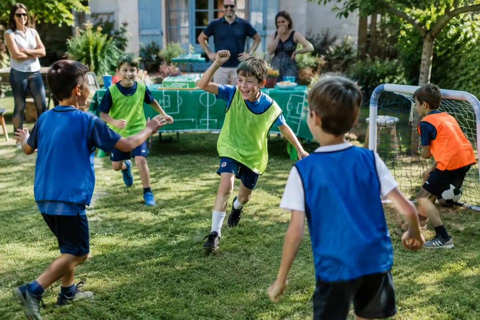 Enfants en chasubles qui célèbrent un but dans un jardin pendant un anniversaire foot, gâteau terrain en fond