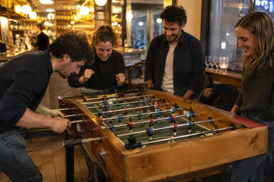 Habitués d'un café de quartier jouant au baby-foot en bois le soir, comptoir en arrière-plan