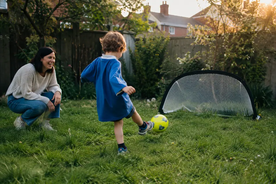 Un enfant de cinq ans frappe un petit ballon vers un mini but dans un jardin, sous l'œil d'un parent