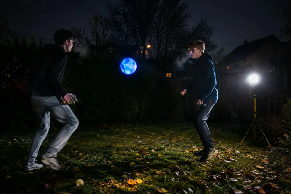Deux adolescents jouent au foot dans un jardin sombre avec un ballon lumineux bleu en l'air