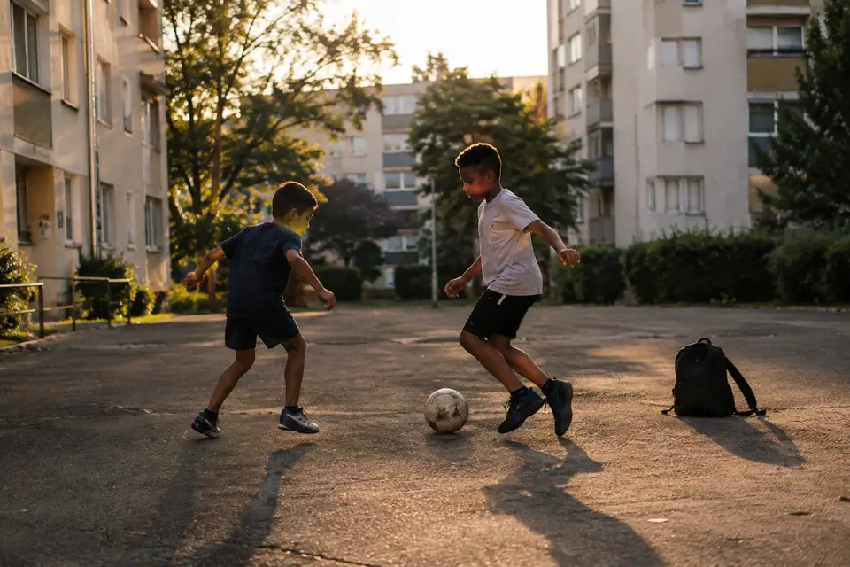 Deux enfants jouent au foot sur un terrain en béton de quartier, un sac à dos comme poteau