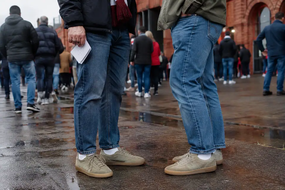 Pieds de deux supporters en baskets basses en daim à semelle gomme et jean droit devant un stade en briques