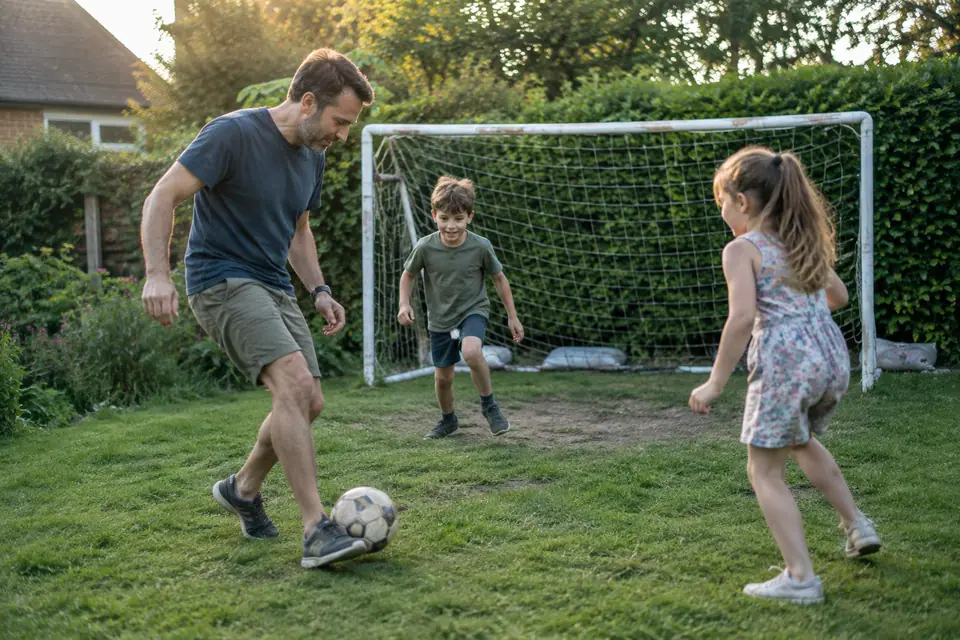 Un père et ses deux enfants jouent au foot devant un but blanc lesté au fond du jardin