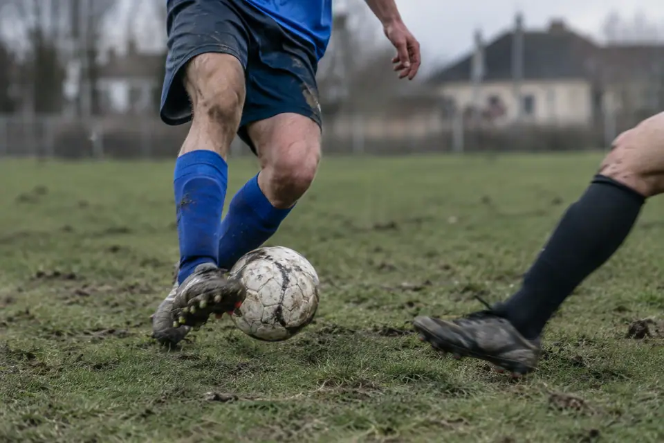 Un milieu de terrain amateur contrôle le ballon sous la pression d'un adversaire sur une pelouse bosselée