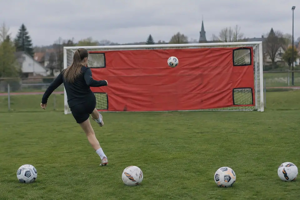 Une bâche cible rouge tendue sur un but de foot, une jeune joueuse frappe vers une ouverture dans l'angle