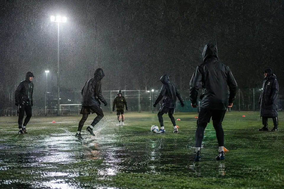 Des joueurs amateurs en vestes de pluie à capuche font un exercice de passes sous une averse nocturne
