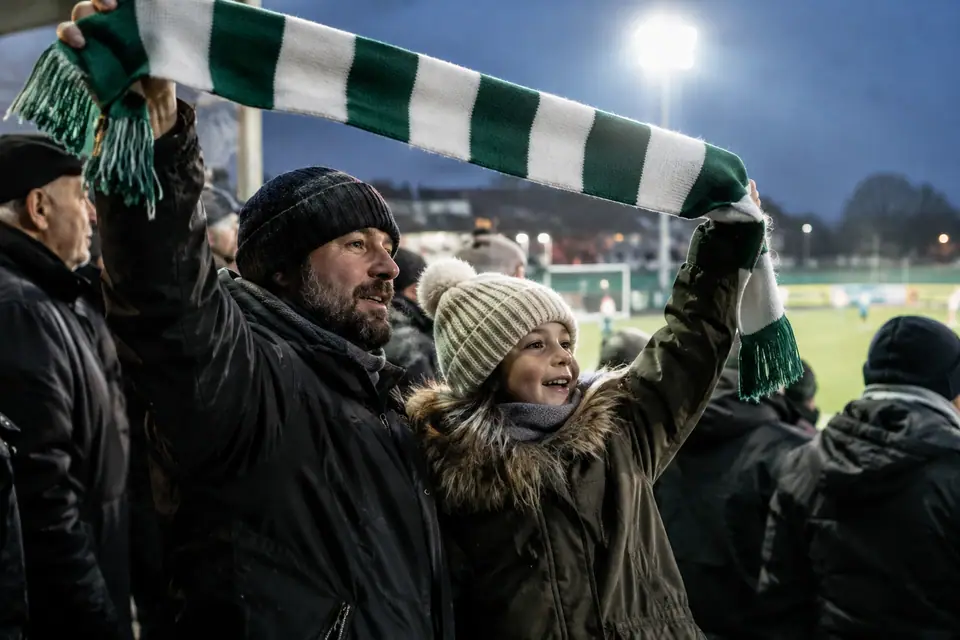 Père et fille levant une écharpe de foot rayée verte et blanche en tribune un soir d'hiver
