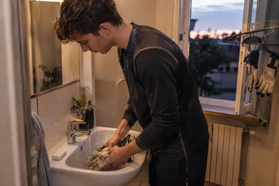 Un jeune gardien rince ses gants boueux dans un lavabo, une seconde paire sèche près de la fenêtre
