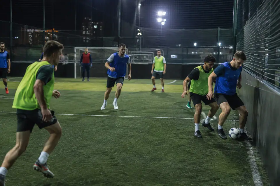 Amis jouant un match de foot à 5 sur un petit terrain synthétique entouré de filets, sous les projecteurs