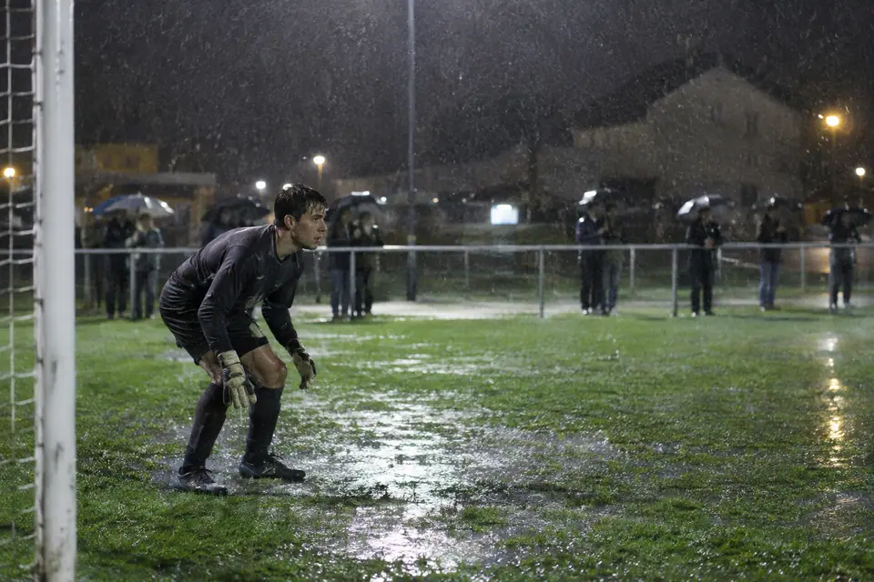 Un gardien amateur accroupi sous une pluie battante, gants ruisselants, sur un terrain détrempé de nuit