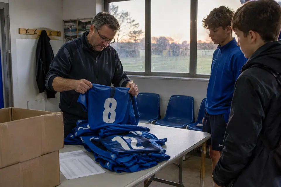 Dirigeant bénévole triant des maillots de foot bleus numérotés sur une table dans le club-house