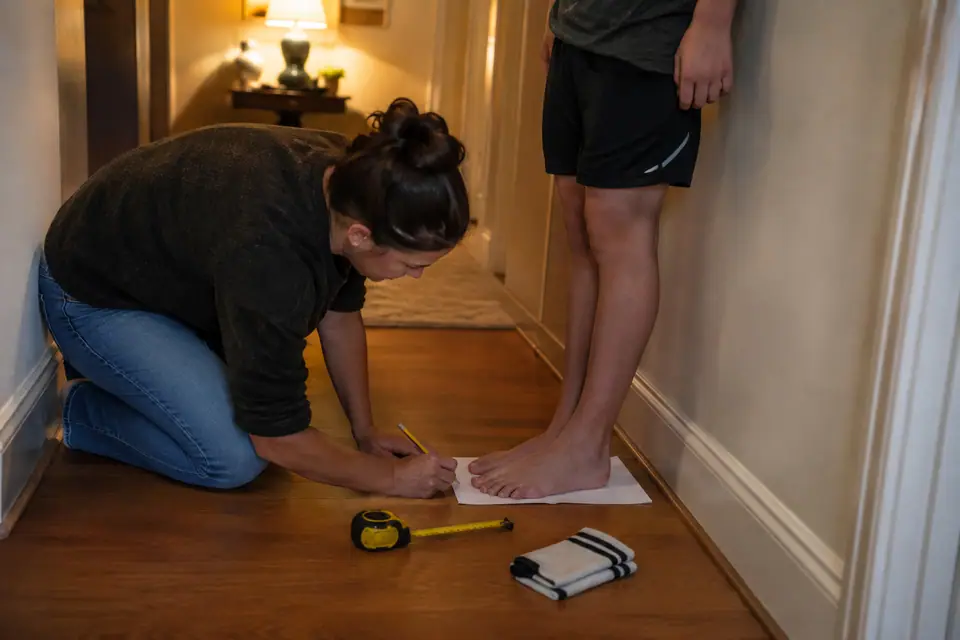 Un parent marque au crayon la longueur du pied d'un adolescent posé sur une feuille contre le mur