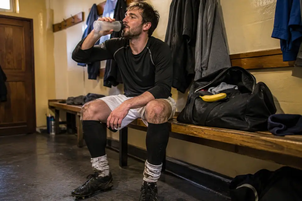 Footballeur amateur fatigué assis dans un vestiaire après le match, manchons de compression aux mollets et gourde à la main