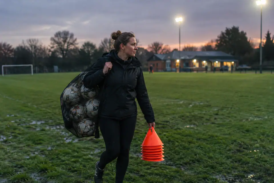 Éducatrice de foot traversant un complexe sportif avec un filet de ballons sur l'épaule au crépuscule