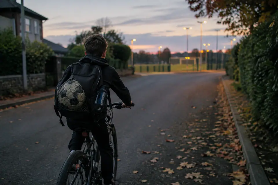 Jeune garçon à vélo avec un sac à dos de sport et un ballon dans un filet, au crépuscule