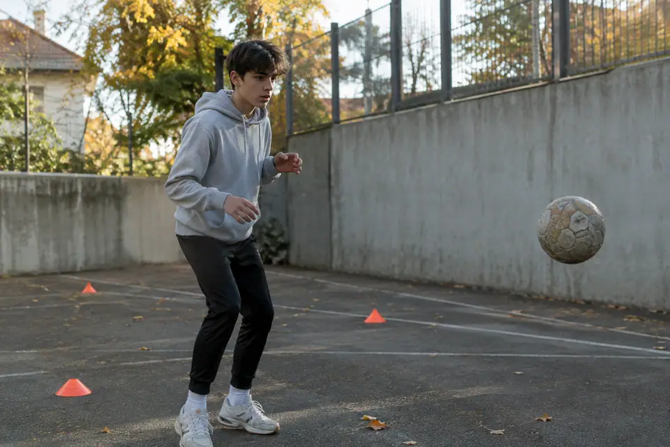 Un adolescent s'entraîne seul au foot contre un mur de city-stade, quelques coupelles orange posées au sol