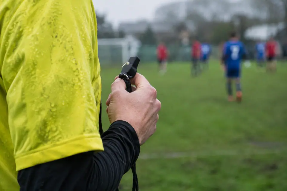 Gros plan sur la main d'un arbitre amateur tenant un sifflet noir sous la pluie, joueurs flous derrière