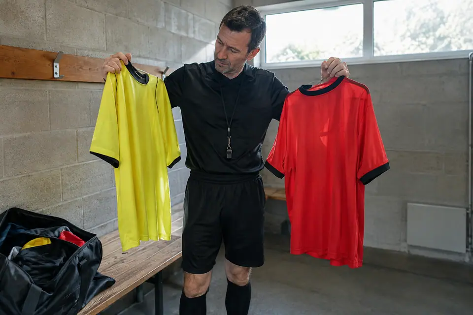 Un arbitre amateur hésite entre un maillot jaune et un maillot rouge dans un petit vestiaire