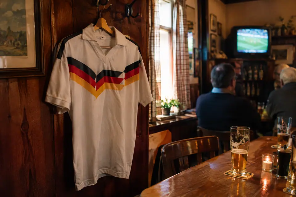 Maillot de foot blanc rétro à bande noire, rouge et or accroché dans un pub