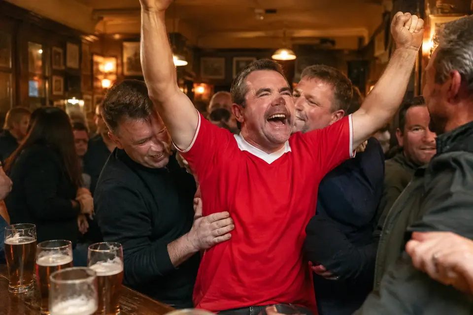 Homme en maillot de foot rouge levant les bras de joie dans un pub bondé, entouré d'amis