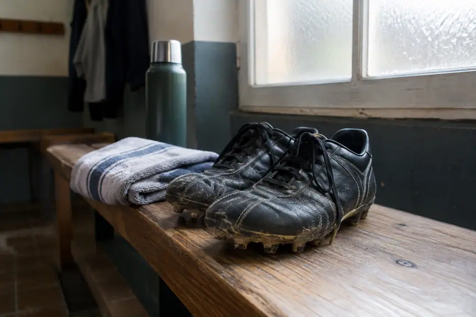 Chaussures de foot en cuir noir boueuses posées sur un banc de vestiaire de club amateur