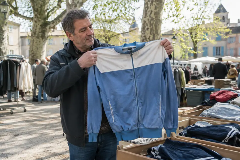 Un homme examine une veste de survêtement rétro bleue et blanche sur un stand de brocante