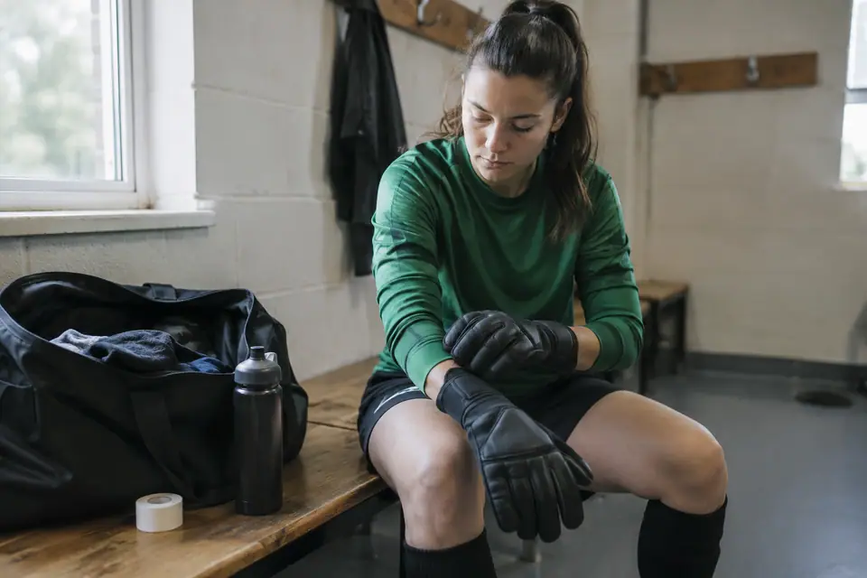 Une gardienne de but enfile ses gants sur un banc de vestiaire avant un match amateur