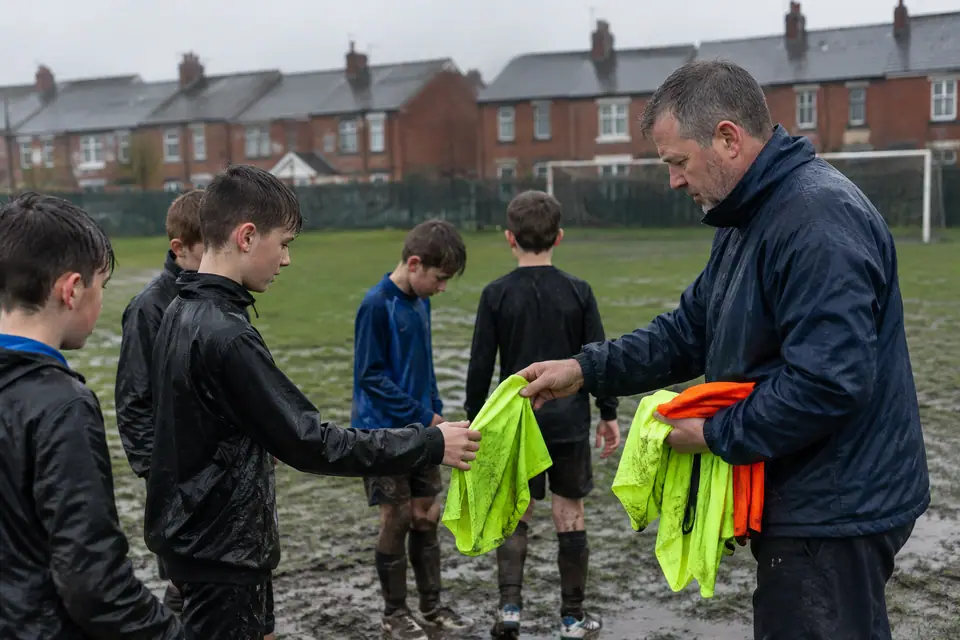 Un éducateur distribue des chasubles à de jeunes joueurs sur un terrain boueux sous la pluie